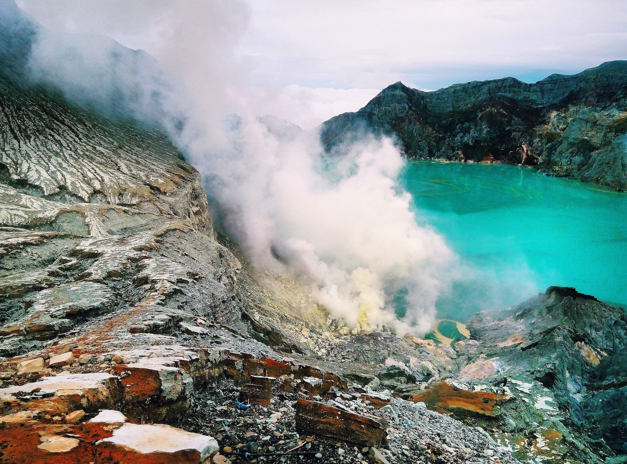 A cloud of volcano smoke with gray volcanic rocks in the foreground and a dramatically teal ocean bay in the background