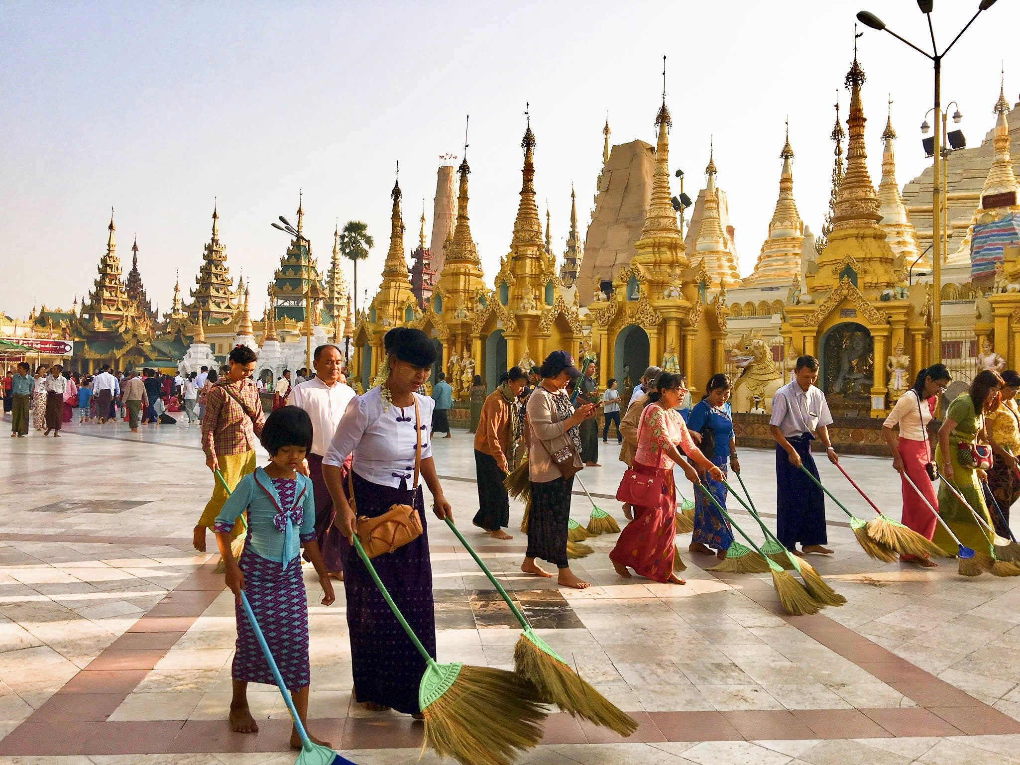 Amidst a crowd, volunteers meticulously sweep the vast marble terrace with large round brooms against a backdrop golden pagodas